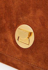 Brown suede handbag with a circular gold metal clasp, featuring embossed branding and a smooth texture. Close-up view highlights material and hardware.
