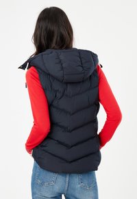 Kvinna med långt mörkt hår som bär en svart huva väst, röd långärmad tröja och blå jeans, med händerna i bakfickorna, vänd bort.