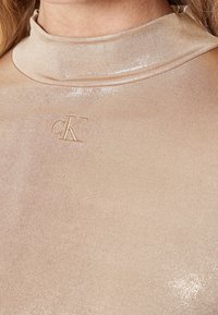 Beige tröja med hög krage i ett blankt, slätt tyg. Har en broderad "cK"-logotyp på framsidan.