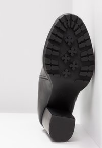 Černá kožená kotníková bota na podpatku s podrobným vzorem podrážky a čtvercovým blokovým podpatkem, umístěná na bílém pozadí.