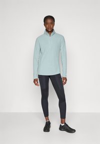 Pullover in pile azzurro chiaro con colletto a mezza zip, abbinato a leggings neri con pannelli testurizzati e scarpe da ginnastica nere.