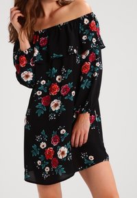 Svart klänning med avskuren axel och volangkrage, med färgglada blommönster i röd, vit och teal. Löst sittande med långa ärmar.