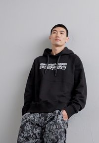 Black hoodie with white text "FEATURE" on the front. Features a kangaroo pocket and drawstrings. Paired with patterned black shorts.