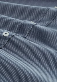 Tejido texturizado en azul oscuro con un sutil patrón de cuadrícula, que presenta botones blancos y detalles de costura visibles.