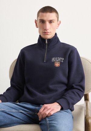 Marineblå fleece-pullover med høj krave, kvart lynlås og broderet logo på brystet. Glat tekstur med ribbede manchetter.