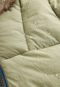 Des gouttes d'eau perlent sur un tissu matelassé vert clair d'une veste résistante à l'eau près d'une fermeture éclair et d'une bordure en fourrure.