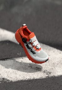 Chaussure de running avec une tige en mesh blanche, des accents orange et un système de laçage BOA. Présente une semelle texturée et un design élégant, flottant au-dessus d'une surface.
