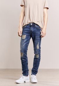 Tummansiniset farkut, joissa on kulutuspisteitä ja haalistunut viimeistely, slim fit -mallilla ja vaaleanvärisellä langalla koristeltuna. Päälle on puettu beige paita.