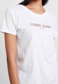 Žena oblečená v bílé bavlněné tričko s potiskem "tommy jeans" v narůžovělé hnědé barvě na hrudi, na jednobarevném pozadí.