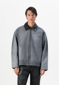 Άνδρας που φοράει υπερμεγέθη γκρι μπουφάν με μαύρο γιακά και μαύρο παντελόνι cargo, στέκεται μπροστά σε λευκό φόντο.