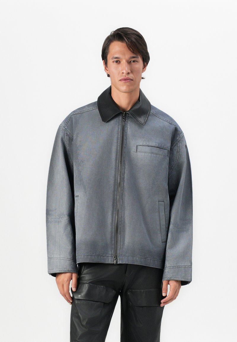 Άνδρας που φοράει υπερμεγέθη γκρι μπουφάν με μαύρο γ�ιακά και μαύρο παντελόνι cargo, στέκεται μπροστά σε λευκό φόντο.