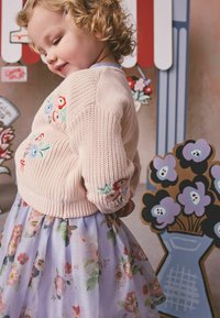 Jauns bērns ar cirtainiem matiem, valkājot rozā izšūtu džemperi un ziedu raksta svārkus, stāvot pie krāsainām apgleznotām ziedu dekorācijām.