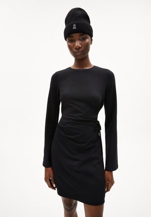 Schwarzes langärmliges Kleid mit einer taillierten Silhouette, seitlichem Bindedetail und weicher Textur. Kombiniert mit einer schwarzen Strickmütze mit kleinem Logo.