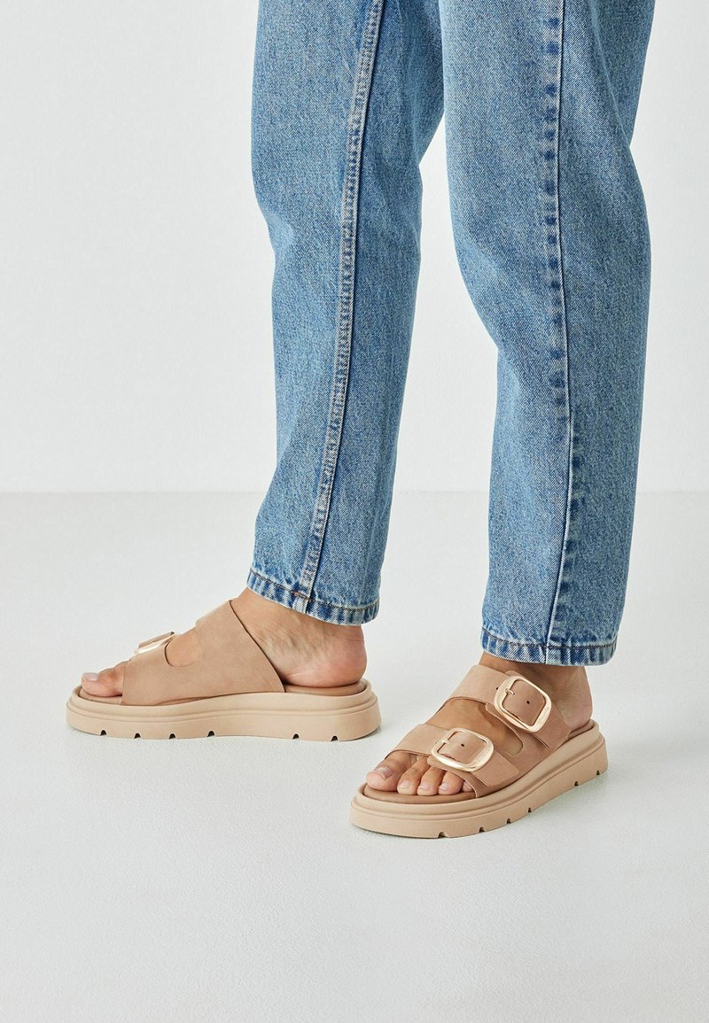 Sandalias de ante beige con dos correas ajustables y una suela gruesa beige, combinadas con jeans de mezclilla azul claro. Diseño plano y abierto en la punta.