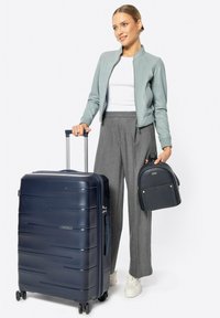 Marineblauwe harde koffer met geribbelde lijnen, uitschuifbare handgreep en wielen. In combinatie met een kleine zwarte rugzak en grijze broek.