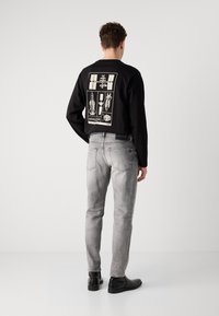 Cămașă neagră cu mâneci lungi și design grafic pe spate, asortată cu blugi gri deschis și cizme negre până la gleznă, evidențiind o ținută casual.