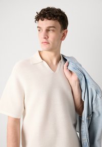 Kortermet, v-hals, kremfarget strikket polo skjorte med teksturerte striper. Lysvasket denimjakke drapert over venstre arm, uten synlig hardvare.