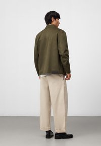 Giacca verde oliva con taglio dritto, tessuto testurizzato e un dettaglio unico sul retro. Pantaloni beige a gamba larga e scarpe nere completano l'outfit.