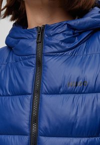 Närbild av en person som bär en blå vadderad jacka med en svart dragkedja och "HUGO"-logotyp på bröstet, som visar kragen och dragkedjans detaljer.