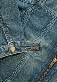 Koptatott világoskék dzseki ezüst színű fém kiegészítőkkel és több látható cipzárral, valamint varrási részletekkel.