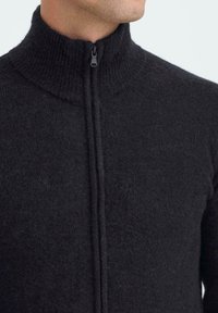 Czarny dzianinowy sweter zapinany na zamek z wysokim kołnierzem, o teksturowanej fakturze i centralnym zapięciu na zamek, podkreślający ciepło i prostotę.