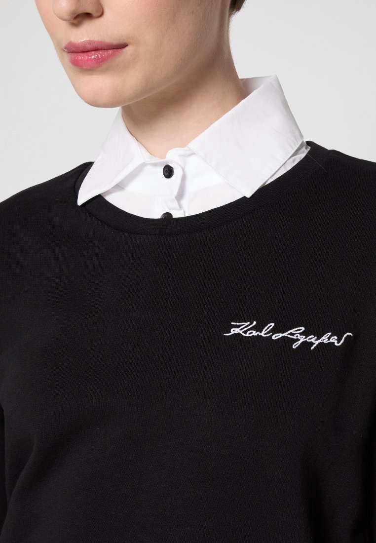 Femme portant un pull noir avec du texte brodé blanc et une chemise blanche à boutons en dessous, avec un col et des épaules raccourcis.