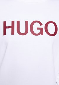 Vit sweatshirt med djärv röd "HUGO"-text. Tillverkad av mjukt tyg med rund halsringning och långa ärmar, som visar en slät textur.