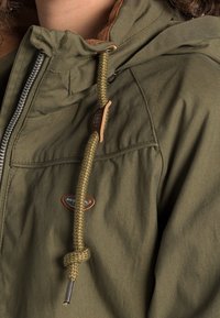 Olivově zelená bunda se světle hnědým límcem. Obsahuje zipové zapínání, texturovanou látku a spletenou šňůrku s dřevěným uzávěrem.