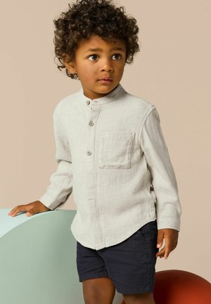 Hellgraues Langarmshirt mit Kragen, drei Knöpfen und Brusttasche. Dunkle Shorts. Kind steht neben geschwungenen pastellfarbenen Formen.