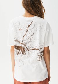 Camiseta de algodón blanca con un diseño gráfico marrón que presenta dos manos y detalles de superficie agrietada en la parte posterior. Ajuste relajado, mangas cortas.