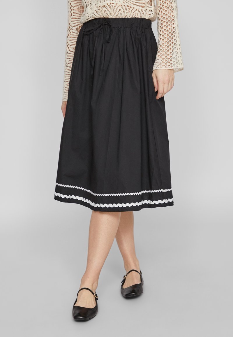 Partie inférieure d'une personne portant une jupe noire jusqu'aux genoux avec bordure blanche en zigzag, un haut en crochet crème et des chaussures Mary Jane noires sur un sol gris.