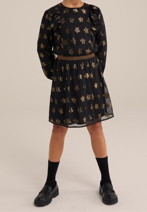 Pige iført en sort kjole med guldstjernemønstre, rynkede skuldre, brun bælte, sorte sokker og sorte loafers, stående på en neutral baggrund.