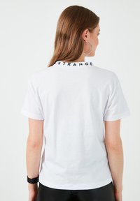 Hvid bomulds T-shirt, kortærmet, med sort "STRANGE" tryk på kraven på bagsiden. Glat tekstur, almindelig pasform.