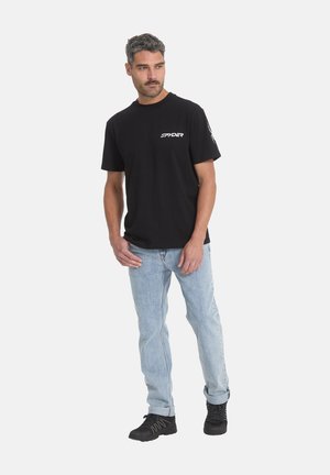 Homme portant un t-shirt Spyder noir, un jean bleu clair et des chaussures noires, debout les mains près des poches, regardant vers la gauche.