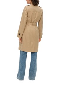 Manteau trench tan avec ceinture amovible et poignets à boutons, associé à un jean large bleu clair, vu de dos.