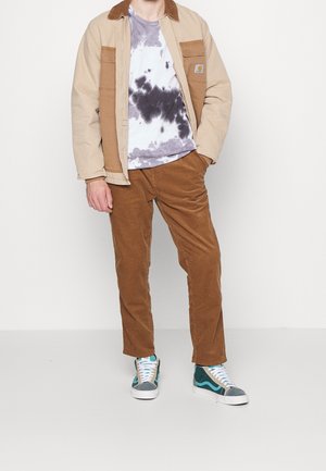 Veste en corduroy beige avec des accents marron, associée à un t-shirt tie-dye gris et violet, et un pantalon en corduroy marron. Baskets bleu et vert.