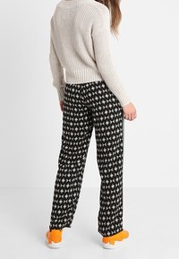 Mujer vestida con un suéter de punto beige, pantalones de pierna ancha de patrón geométrico negro y zapatillas brillantes naranjas, de pie frente a un fondo blanco sencillo.