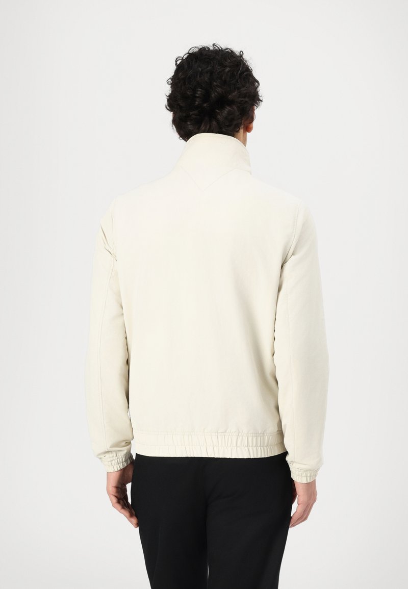 Chaqueta beige clara con un collar alto, puños y bajo elásticos, y una textura suave. No tiene patrones visibles ni detalles de hardware.