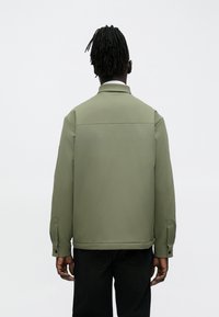 Olive green jacket with a structured design, buttoned cuffs, and a back yoke. Made from smooth fabric, features no visible patterns or hardware.