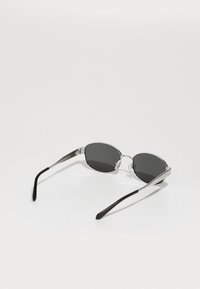Ochelari de soare cu rame metalizate argintii, lentile gri, și vârfuri negre ale brațelor, având o formă ovală și un design minimalist.