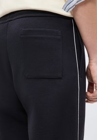 Κοντινό πλάνο ατόμου που φοράει μαύρο υφασματόπαντελόνι με πίσω τσέπη και λεπτή λευκή πλευρική ρίγα, σε συνδυασμό με κρεμ πουκάμισο και ριγωτό μανίκι.