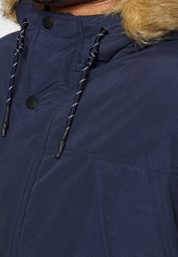 Mörkblå jacka med foder av fuskpäls i huvan. Har svarta tryckknappar och justerbara snoddar. Slät yta med en något matt finish.