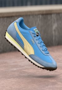 Sapatilhas Puma com um upper sintético azul, detalhes em amarelo claro, materiais de camurça suave, sola de borracha texturizada e design de atacadores.