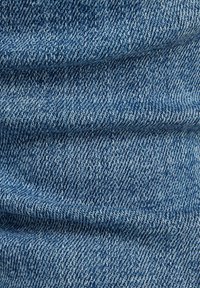 Tejido de mezclilla azul con un tejido de sarga clásico, que presenta un lavado medio. La textura es suave, con costuras visibles y sutiles variaciones en el color.