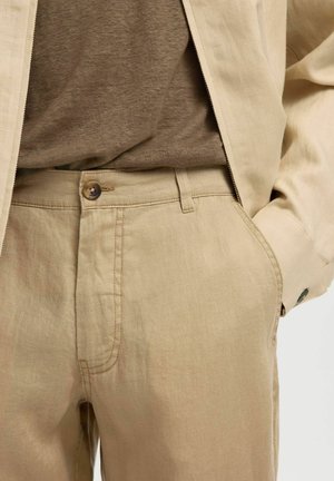 Beige linbukser med knapp og glidelås, kombinert med en matchende beige jakke og en brun skjorte stukket inn, med hånden i lommen.