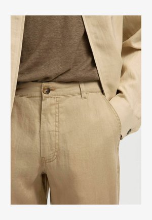 Beige linbukser med knapp og glidelås, kombinert med en matchende beige jakke og en brun skjorte stukket inn, med hånden i lommen.