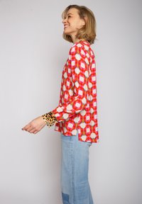 Langärmlige Bluse mit einem roten, rosa und weißen kreisförmigen Muster. Die Ärmel haben einen Leopardenmuster-Bündchen. Leichter, glatter Stoff.