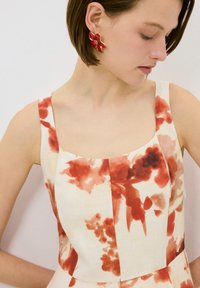 Un vestido sin mangas y ajustado de tela ligera con un patrón floral rojo. Combinado con pendientes en forma de flor rojos con acentos dorados.