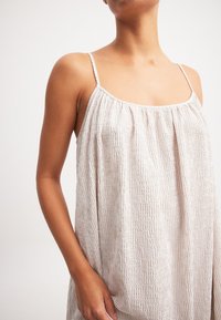 Šviesiai smėlinė, be rankovių suknelė su plonomis petnešėlėmis, pagaminta iš tekstūruoto audinio su subtiliu raukinių raštu, laisva siluetu ir kvadratiniu iškirpimu.