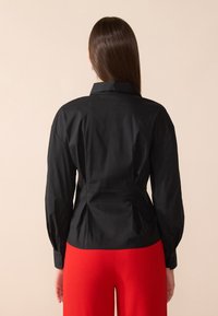 Maglietta nera a maniche lunghe con colletto strutturato, pieghe sul retro per dare forma e polsini con bottoni, abbinata a pantaloni larghi rossi.